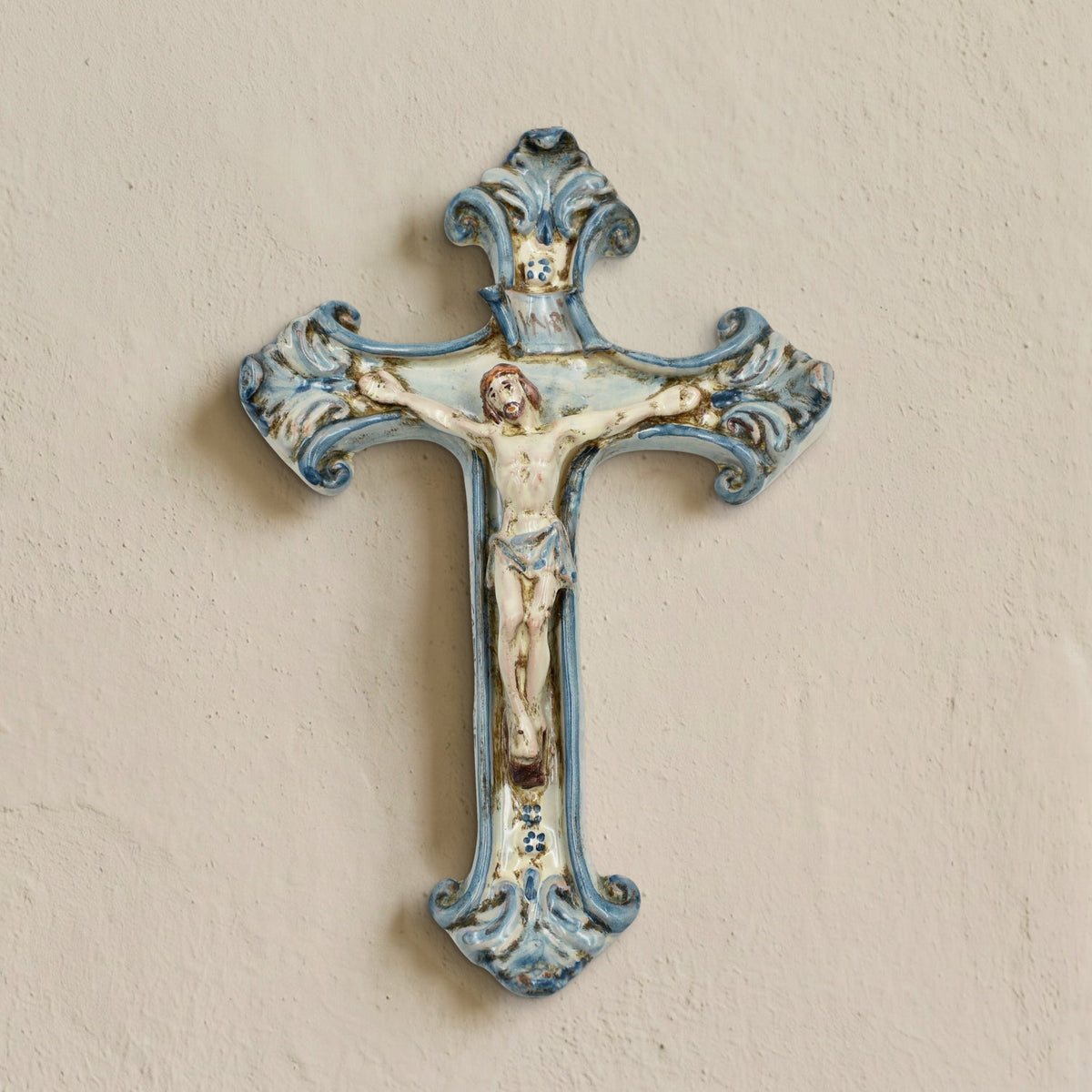 Crucifix en céramique de Caltagirone Mono Bleu accroché à un mur clair, fait main avec Christ en relief et volutes peintes en bleu