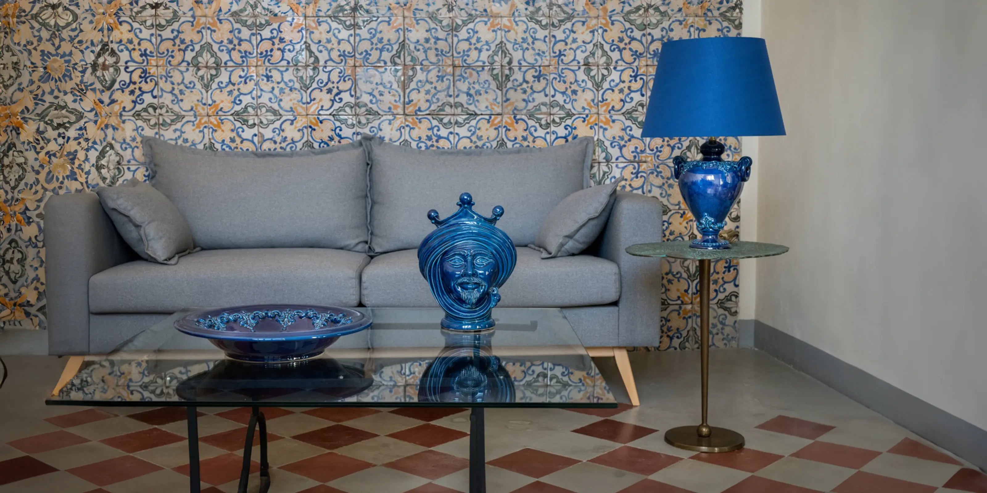 Interno di una casa arredata con le Ceramiche di Caltagirone Sofia in colorazione Blu Intenso, sul tavolo un piatto in ceramica e a fianco una Testa di Moro alta 30 cm, a destra su un tavolo troviamo un lume con un paralume in colorazione blu.