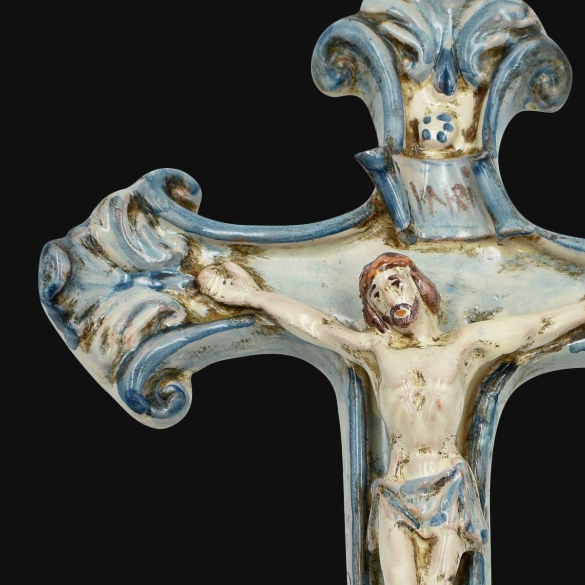Detalle del brazo izquierdo del crucifijo de cerámica de Caltagirone Mono Azul, con volutas en relieve pintadas a mano en azul y la figura de Cristo